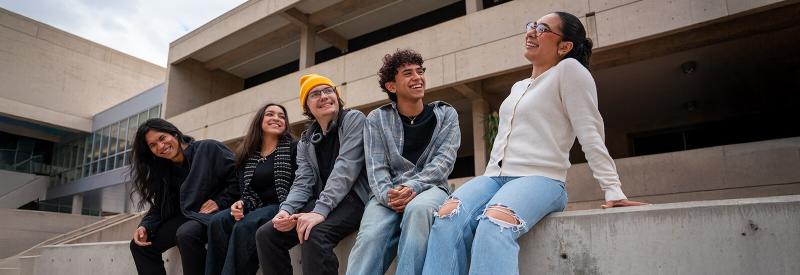 5 students sit on a pony wall in a west campus courtyard